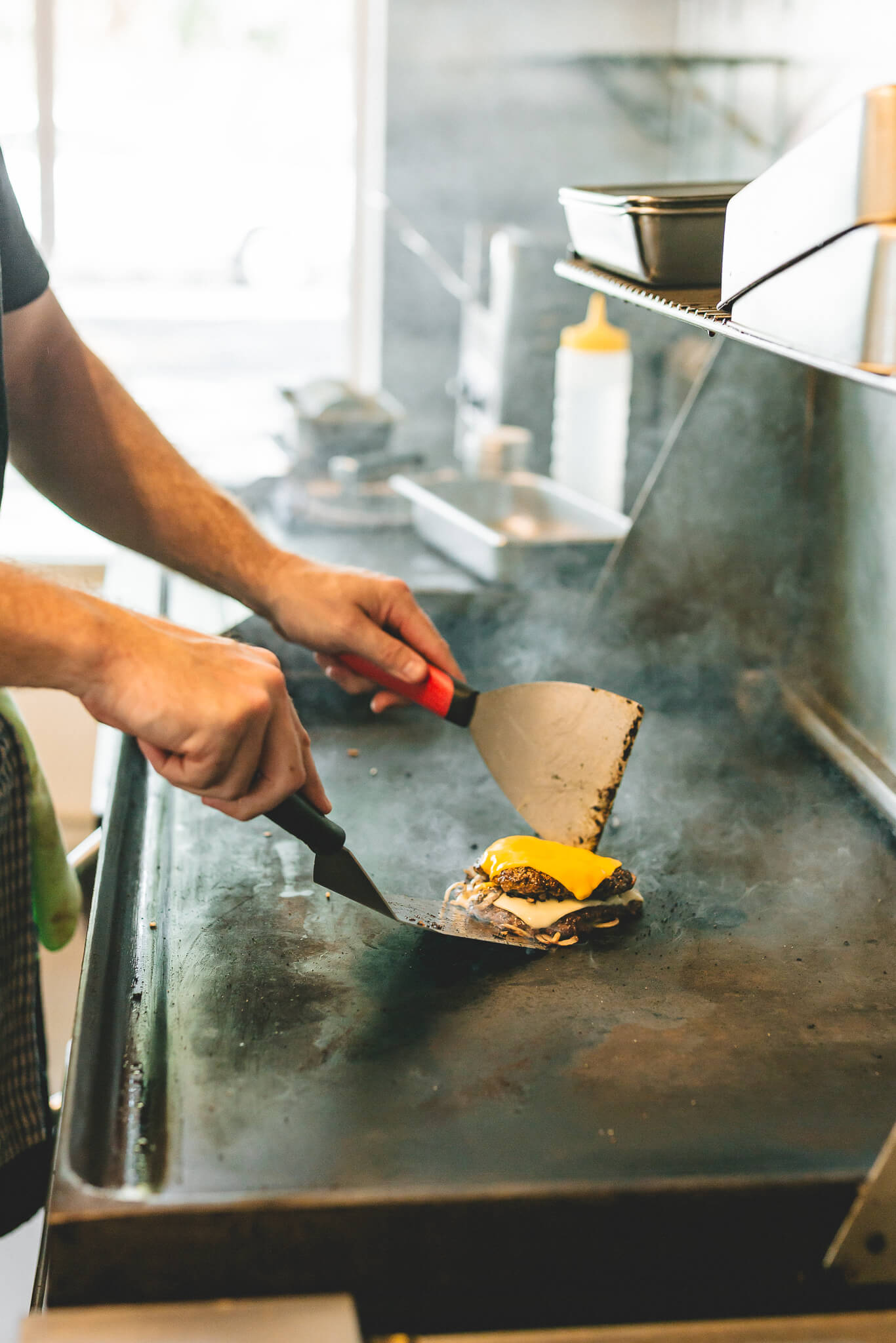 A cook smashing a cheese-topped patty on the flat-top griddle, smoke rising.