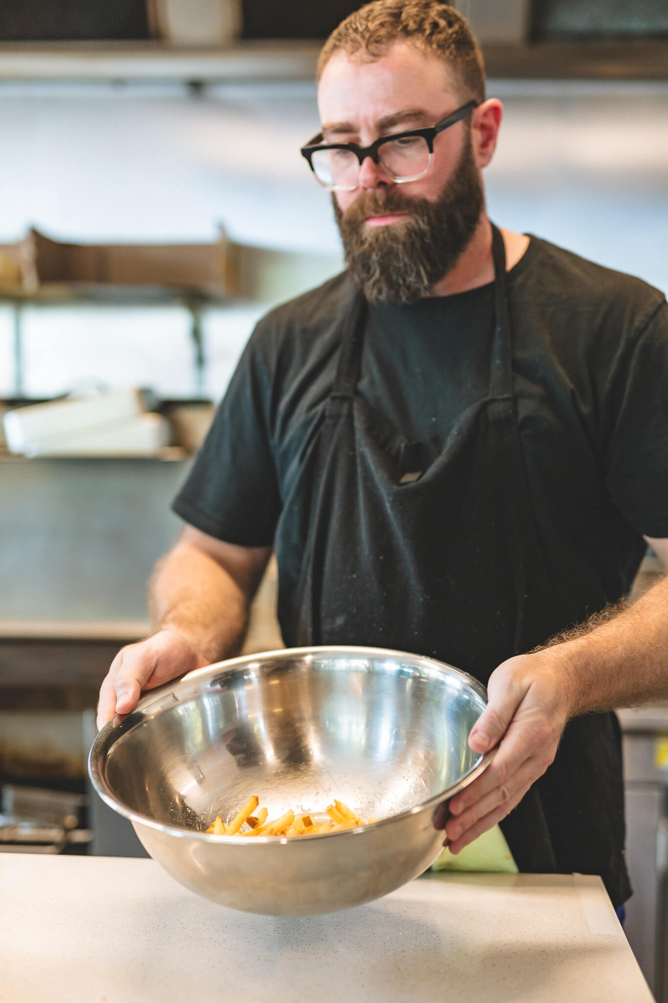 A cook in an apron carrying a steel bowl of freshly fried chips through the kitchen.